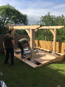 carpenters installing green oak timber framed pergola onto granite staddlestones in a sunny garden